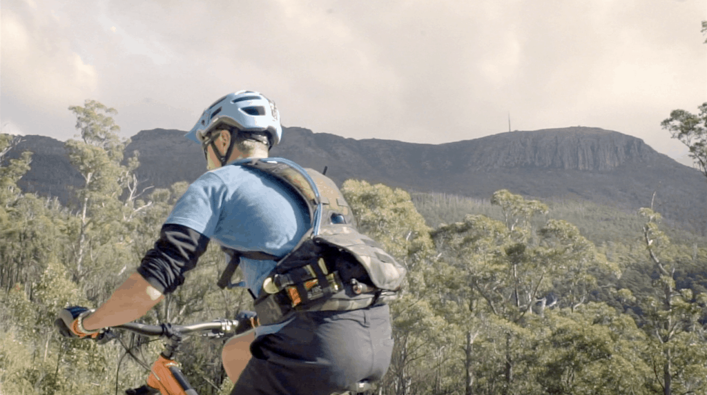 A man cycling with an Enduro Pack on his back, Mount Wellington in the background.